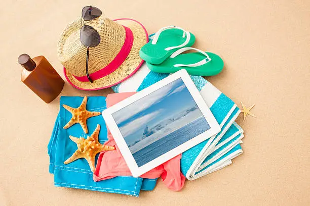 Beach accessories on beach- sunglasses, towel, starfish, sunscreen, straw hat,, swimsuit and pc tablet on the towel.