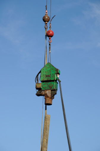 A pile driver being lowered onto a wood pole by a steel cable attached to a crane and stabilized by 2 wrecking balls. A cloth strap is attached to the pole and one of the balls.
