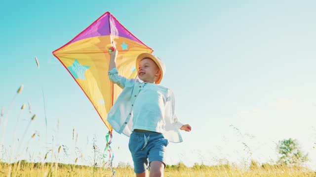 Fast little boy runs across the field with a kite in his hands fluttering in wind over his head. Front view on child walks in nature in summer and has fun play. Family trip out of town to fresh air.