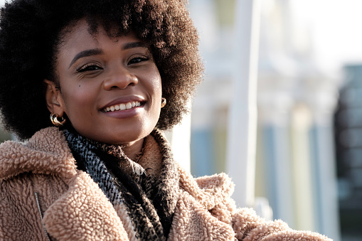 Smiley young black curly woman looking at camera outdoors. kindness and sympathy concept. Warm winter light in a cold morning.