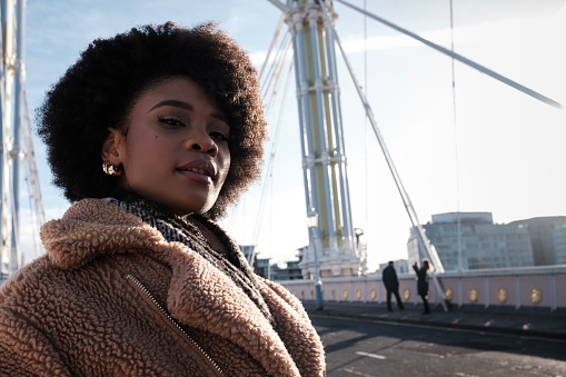 Portrait of young curly black woman posing on historical bridge in London. She is looking at camera and she is wearing winter outfit. Two blurry people taking pictures on the other side of bridge.