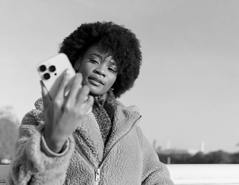 Black and white portrait of young curly black woman doing selfie outdoors. It is a cold winter day and she is wearing coat. Image made with analog camera. Old and new technologies living together