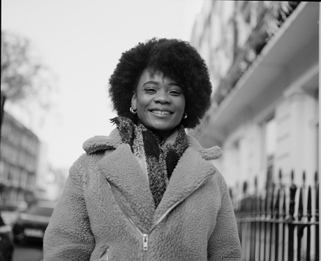 Monochromatic portrait of happy young black curly woman posing in a London street. It is a cold winter morning and she wearing a thick coat and scarf. Portrait made with analog medium format camera.