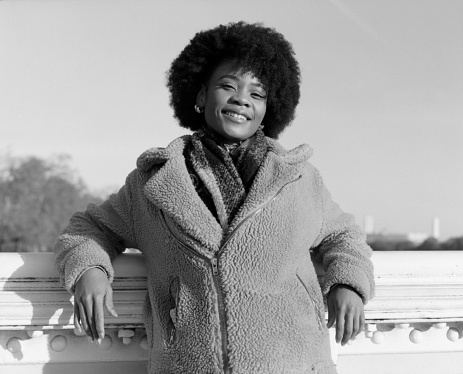 Black and whiter portrait of happy young black woman leaning on bridge banister. She is enjoying the winter sun and she is wearing and thick coat and scarf. Image made with analog camera.