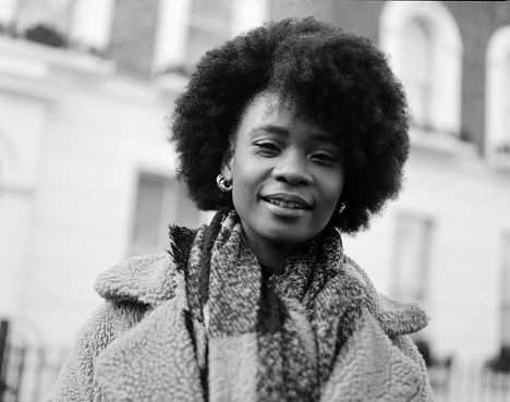 Analog portrait of smiling young curly black woman looking straight to camera in the street. She has a building behind and she is wearing winter outfit. Image made with medium format analog camera.