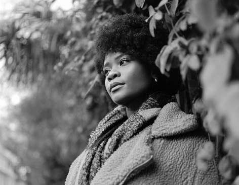 Analog portrait of pensive young black curly woman posing between brushes. She is wearing a winter coat and scarf. Nostalgic image made with a film medium format camera.