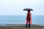 A woman with an umbrella in a red raincoat stands in the rain on the seashore. Rainy weather at sea.