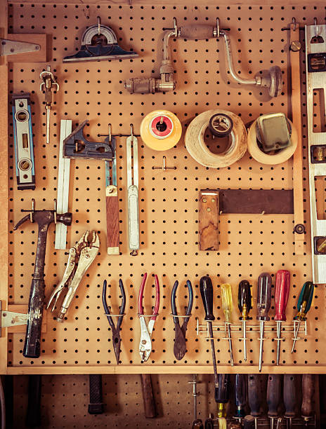 Woodworking Tools Hanging In Workshop. The background is old yellowed pegboard. There is an assortment of screwdrivers, pliers, wire cutters and tapes on the wall. A hammer and a square and two levels are also present. There are no people in the image. 