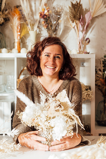 Smiling middle aged female florist with short hair looking at camera while sitting at desk with hands around arranged dry flowers bouquet in floristry shop against shelves in daylight