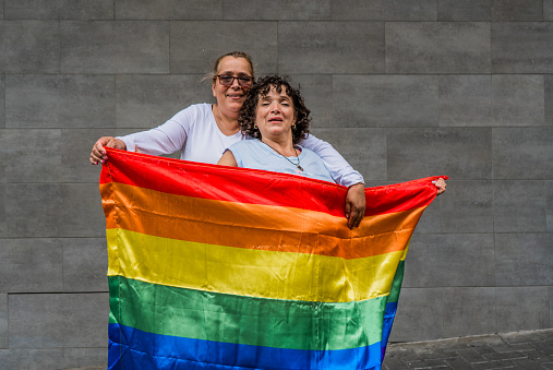 portrait of a couple of women embracing outdoors and smiling as they smile and hold the LGBTIQ+ flag.