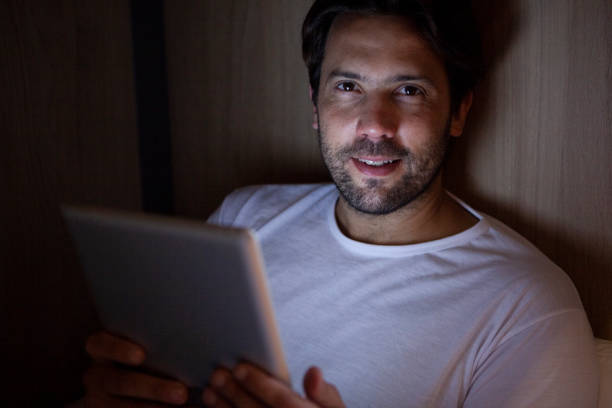Portrait of adult man looking at the camera while holding digital tablet lying on bed