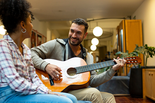 Mid shot of young adult man playing guitar while sitting on sofa with friend
