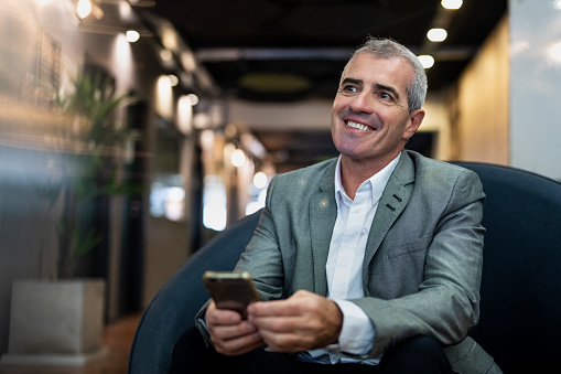 Front view portrait of senior businessman sitting on chair while using smart phone