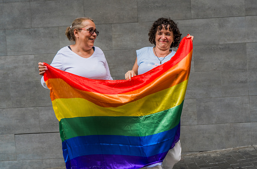 a pair of two women smiling while holding the gay flag in their hands