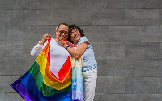 portrait of a beautiful couple of adult gay women embracing while holding the LGBTIQ+ flag in their hands.
