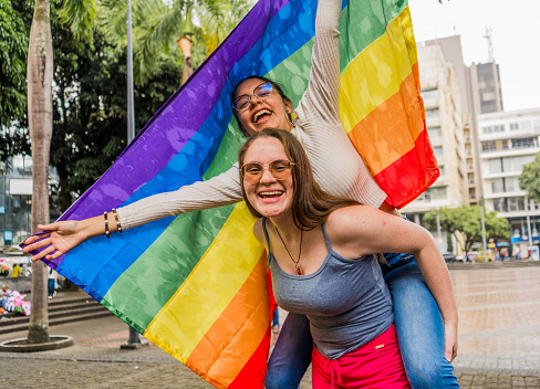 women playing in the park riding on top of each other while smiling with their hands open holding the gay flag.