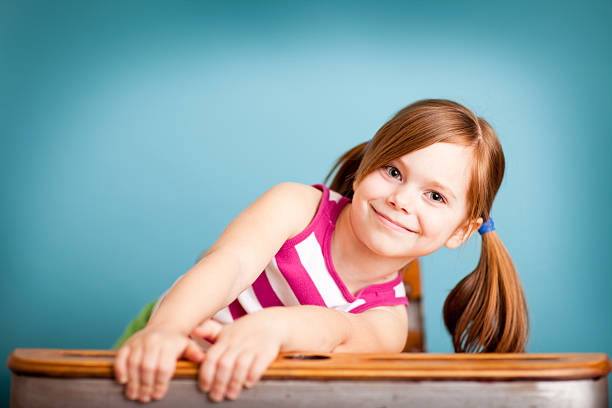 Color photo of a young girl student smiling while sitting in a school desk on blue background.