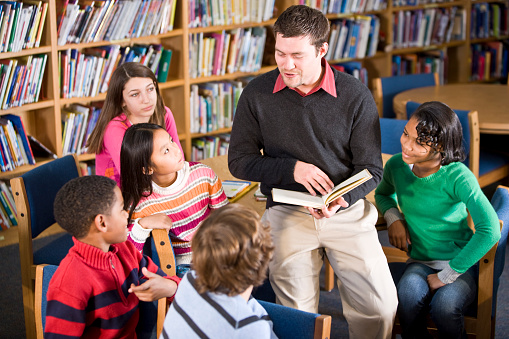 Young man in library with group of students, 9 to 11 years.  Shallow DOF, focus on man and Asian girl.