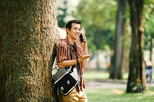 Smiling young man leaning on tree when talking on phone with friend