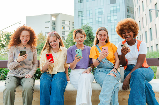 Multiracial group of young female friends enjoying and smiling using their mobile phone app sitting at downtown. Diverse teenage girls having fun sharing messages with each other on cellphone