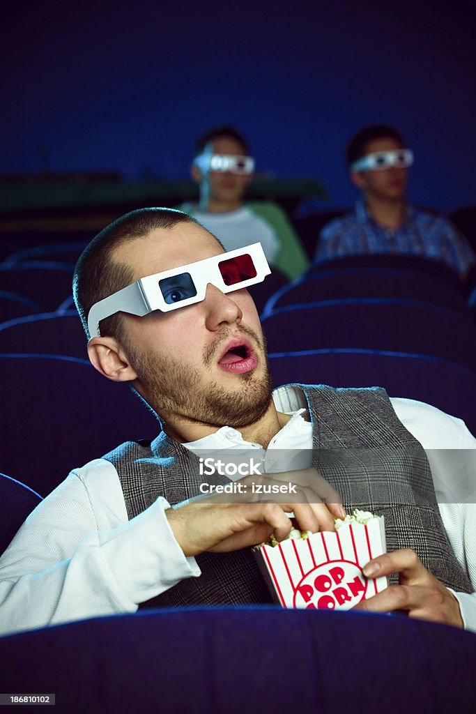 Young man in 3D move theater "Young adult man wearing 3D glasses, sitting in the cinema, watching scary 3D move and eating popcorn. He looks very frightened." 3-D Glasses Stock Photo