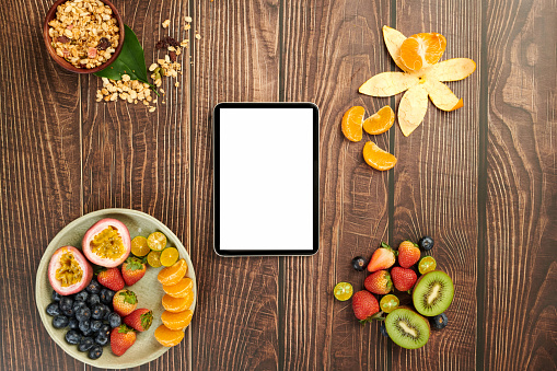 Tablet computer on wooden table next to plate with fresh fruits and granola bowl
