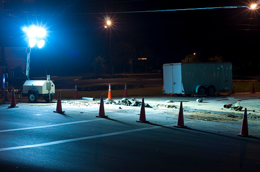 A bright light illuminates a road which has a large section of it torn out - traffic cones block the area off.