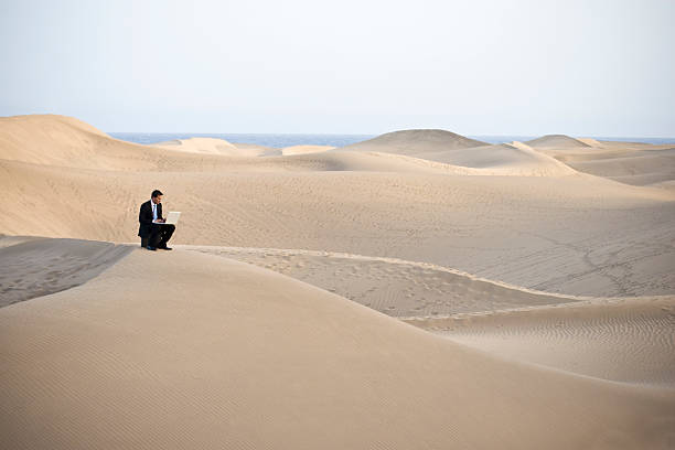 Man working on laptop sitting on a sandy desert hill stock photo