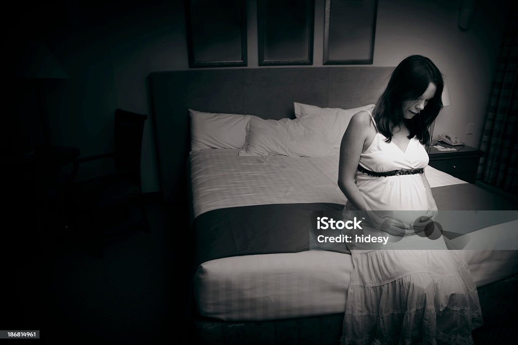 A black and white photo of a pregnant woman sat on the bed Young mother to be sits alone and sad in a hotel room.  Monochrome image. Pregnant Stock Photo