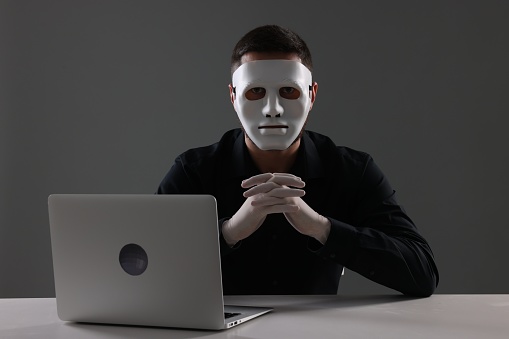 Man in mask and gloves sitting with laptop at white table against grey background