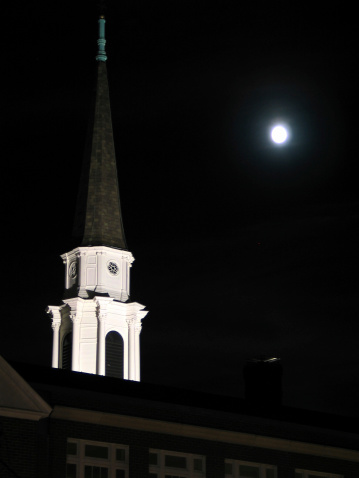 A shot of a lighted steeple from below with the moon in the background.