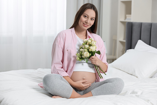 Beautiful pregnant woman with bouquet of roses in bedroom