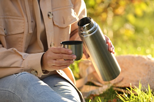 Picnic time. Woman with thermos and cup lid on green grass outdoors, closeup