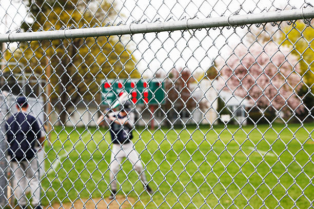 Baseball Player Taking Warmup Swings Beyond Chainlink Fence Beyond a sturdy protective chainlink fence safety backstop, a high school baseball player takes warmup swings before going up to bat in a baseball game. Selective focus on the fence - with the player, coach and far background scoreboard intentionally blurred. high school baseball stock pictures, royalty-free photos & images