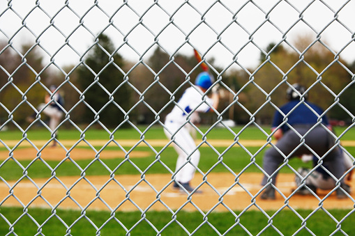 Beyond a sturdy protective chainlink fence safety backstop, a baseball pitcher is winding up to pitch to a waiting batter, catcher and umpire during a high school baseball game. Selective focus on the fence - with the players and umpire intentionally blurred.