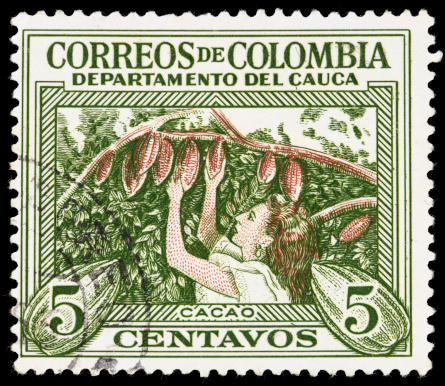 A vintage Colombian postage stamp containing an illustration of a woman picking cocoa pods from a cocoa plant.