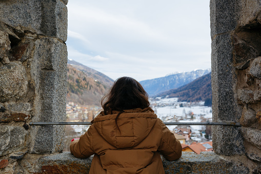 rear view of standing girl in warm jacket watching snowy mountains from the big window