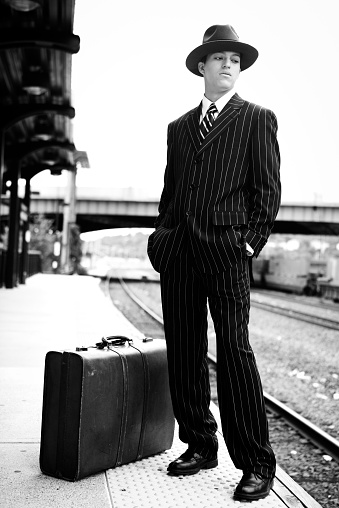 A man in 1940's style clothing standing on a train platform with a suitcase.