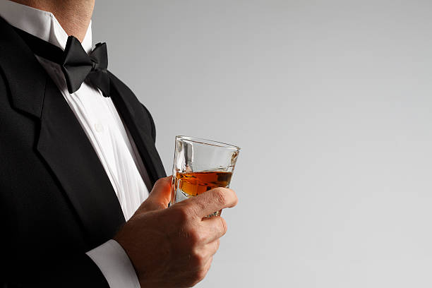 homem de smoking segurando captura de whiskey vidro contra fundo cinza - high society men tuxedo party imagens e fotografias de stock