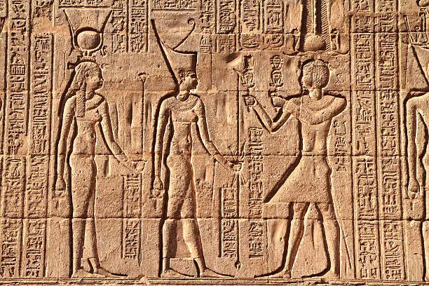 "Sunk relief on the east facade of the Hypostyle Hall of the Temple of Khnum, Esna, Egypt.  The relief depict the pharaoh (on the right) making an offering to the god Khnum (also Chnum, Knum, Khnemu) in the centre.  The pharaoh wears the double ostrich feather Shuti crown.  Khnum appears in human form and wears the red crown, or Deshret, of lower Egypt .  He carries the ankh, symbol of life in his right hand.  The figure on the left is probably Hathor, goddess of love music and dance.  She wears the solar disk and cow horns.  The Ptolemaic and Roman temple was dedicated to the god Khnum.  Only the hypostyle hall has survived and is now located in the centre of Esna."