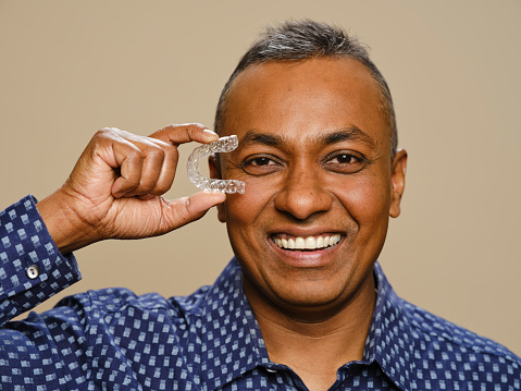 A mature Indian ethnicity man, holding clear dental teeth aligners.