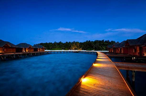Blue Maledives - Fihalhohi Island Resort overwater bungalows boardwalk with view of an island of the Maldives to the blue hour water bungalow maldives stock pictures, royalty-free photos & images