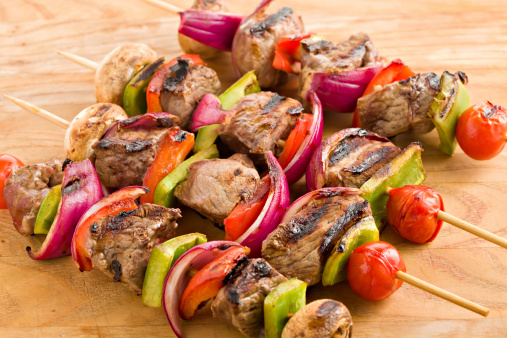 An overhead close up view of a wooden cutting board with four sirloin and vegetable shish kabobs fresh off the grill.