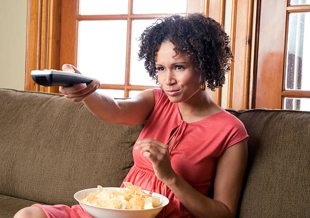 Woman eating chips and pointing remote watching television stock photo