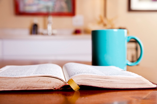 Open bible on kitchen table with coffee mug.  Selective focus on the front edge of the Bible.    