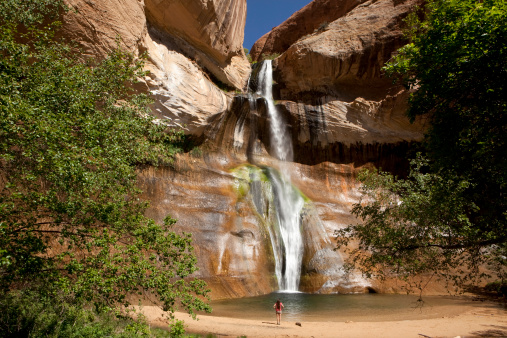 A woman stands in front of the 126 feet high Calf Creek Falls as it cuts through the Navajo Sandstone in Grand Staircase Escalante National Monument in Utah. The high desert of Calf Creek Canyon has been inhabited by early Native American ancient puebloans and early ranching pioneers. Box elder trees and gambel oak blow in the wind as mist drifts off the falls.