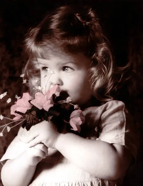 Little girl holding flowers by her face. Eyes have been hand-touched with blue and flowers touched with pink. In sepia.
