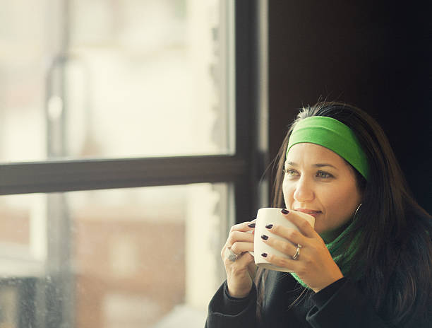 drinking coffee by the window mid adult hispanic woman drinking warm beverage by a huge window on a winter snowy day. ca04 stock pictures, royalty-free photos & images