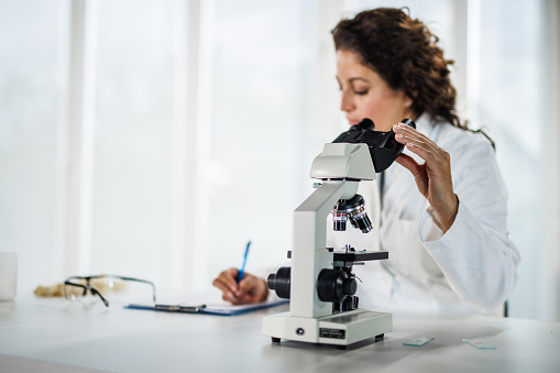 Copy space shot of focused mid adult female scientist writing results on the clipboard while analyzing scientific samples under a microscope when conducting research at the laboratory.