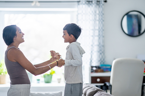 A little boy dances with his Mother around the living room on a casual Saturday afternoon.  They are both dressed comfortably and are smiling as they enjoy spending time together.
